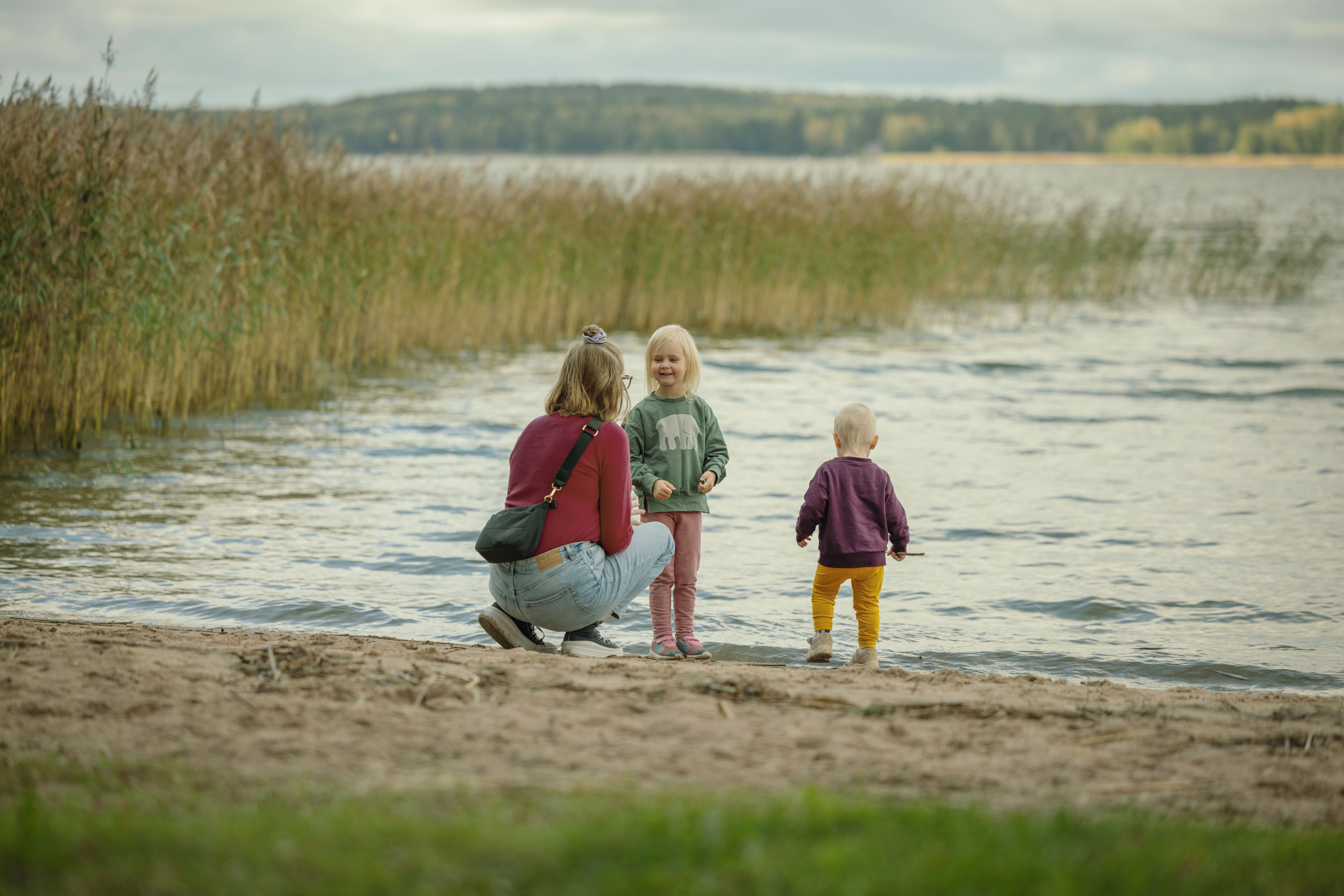 Äiti ja lapset uimarannalla heittelemässä kiviä mereen.